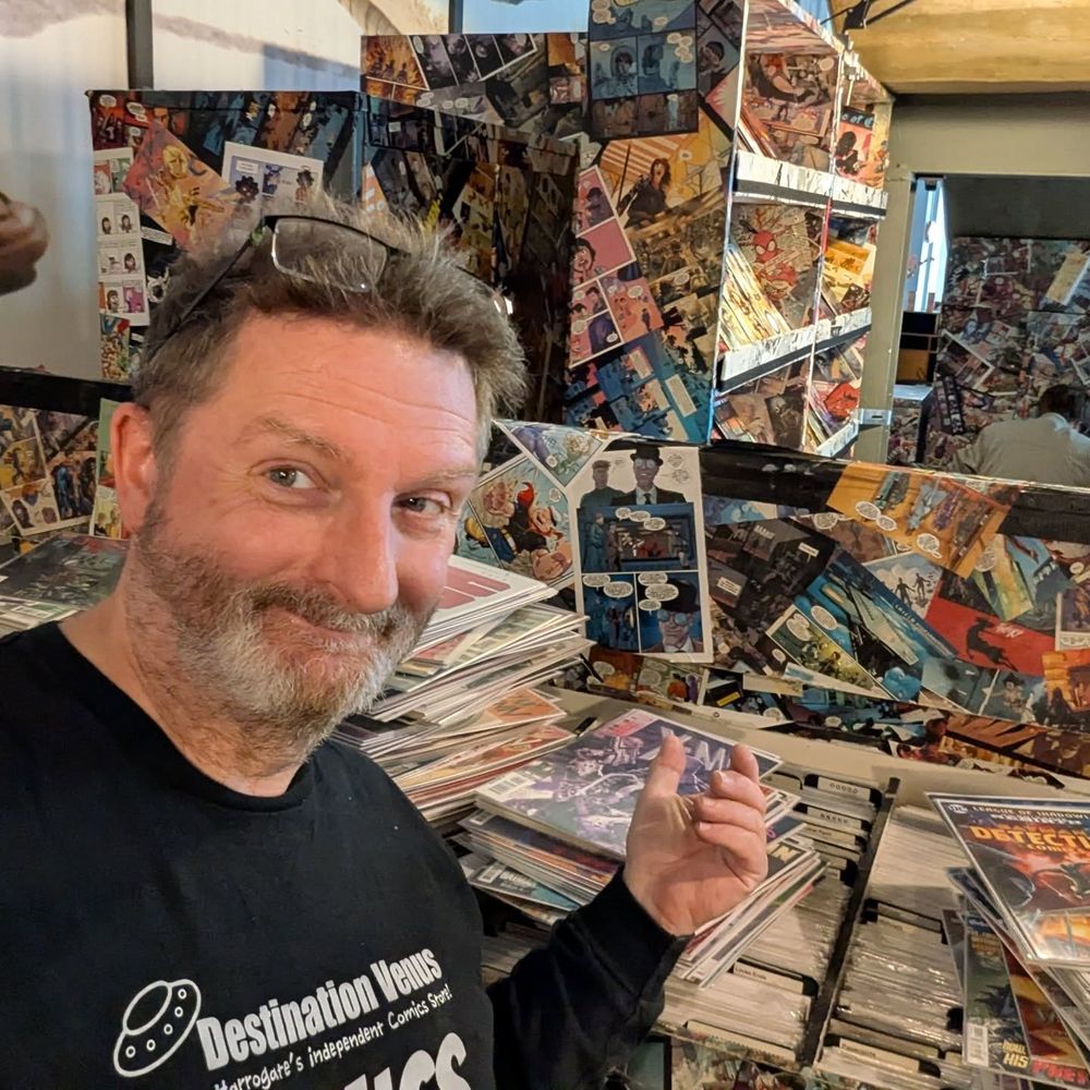 Regie looks at the camera, motioning to the back issue section behind him. It is strewn with bagged and boarded comics waiting to be sorted and filed for customers to browse. Behind that you can see the rest of the store in all its decopaged glory.