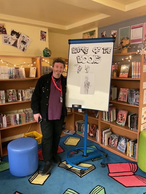 Regie, wearing black jeans, black jacket and a purple shirt - possibly the most colourful outfit he has ever worn in public - stands next to a whiteboard bearing the legend "Battle of the Books" with bookshelves in the background. The carpet is also decorated with books. There might be a theme developing here...
