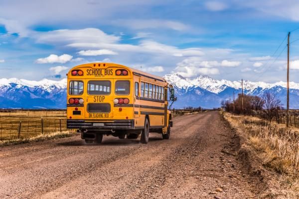A school bus drives down a rural dirt road. A mountain range can be seen in the distance.