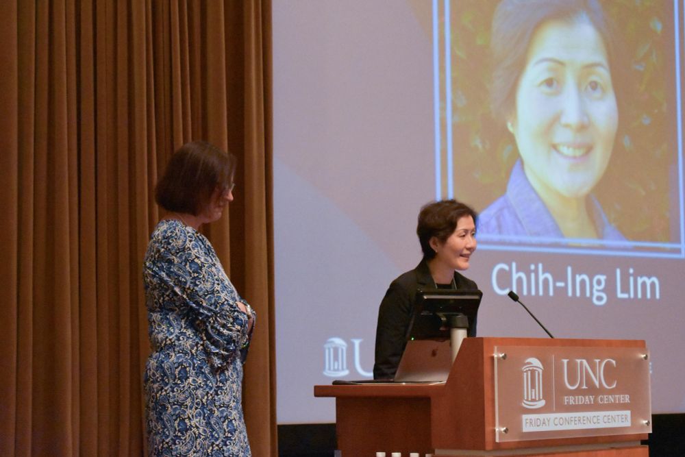 Chih-Ing Lim stands at a lectern in front of a presentation screen and addresses the attendees of the Inclusion Institute. Paula Grubbs stands to her left. 