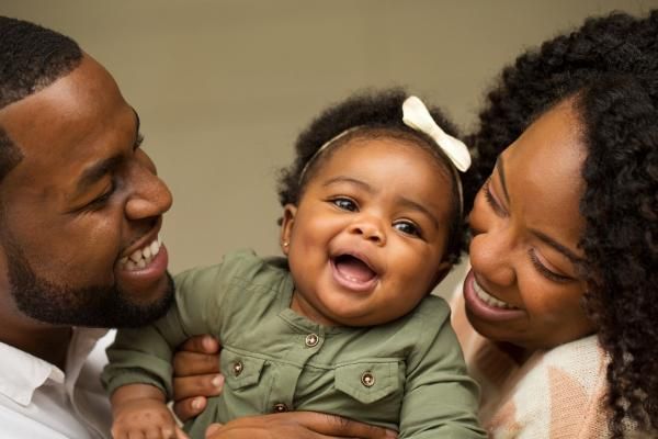 A Black family consisting of a mom, dad, and a toddler-aged daughter smile with their faces close together.