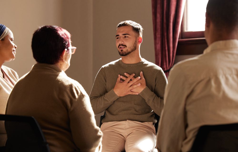 A man with short hair and a beard holds his hands to his chest while addressing a support group