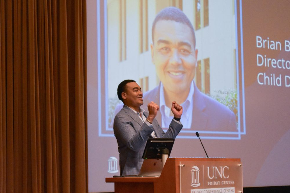 Brian Boyd stands at a lectern and holds his hands up in celebration while addressing the Inclusion Institute attendees.