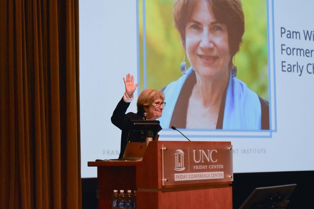 Pam Winton stands at a lectern in front of a presentation screen and waves to the attendees of the Inclusion Institute.