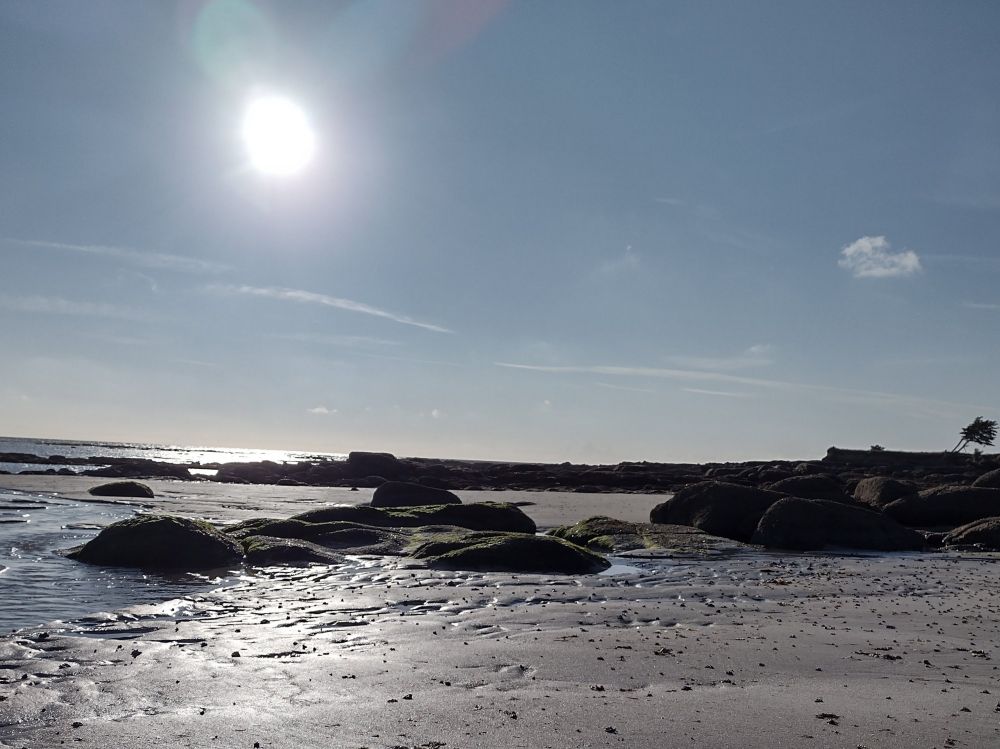 Grey sandy beach with rocks and sea, with sun shining on the sand and the sea