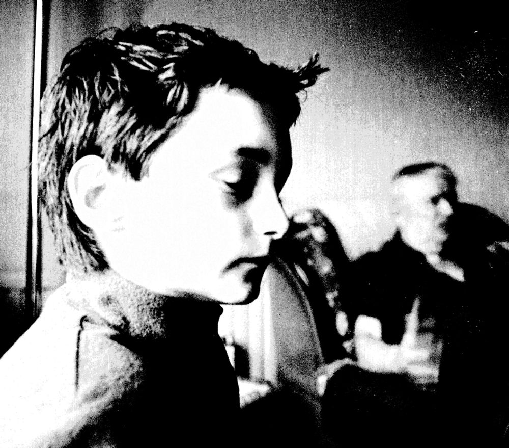 black and white grainy image of young pson in the foreground with quiff with older man in the background in an armchair
