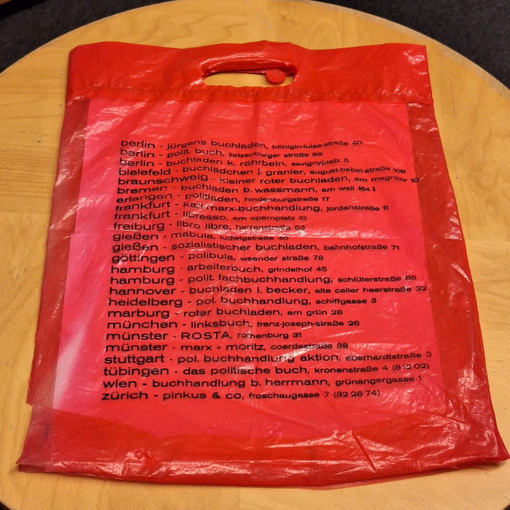 Rote Plastiktüte (Rückseite), darauf in Schwarz gedruckt die Namen diverser linker und alternativer Buchhandlungen in Deutschland, Österreich und der Schweiz der 1970er und 1980er Jahre.
