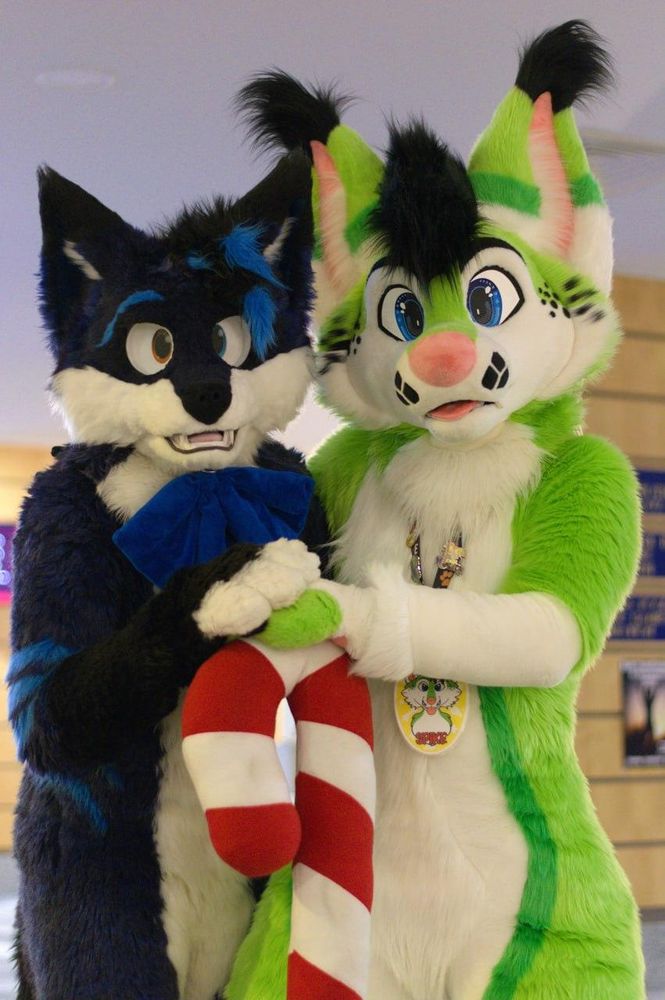 A photography showing the upper body of two fursuiter, standing side by side and looking directly into the camera while both of them having one of their paws placed on top of a huge plush candy cane, which is standing on the ground in front of them. The background vaguely shows the lobby of a hotel.