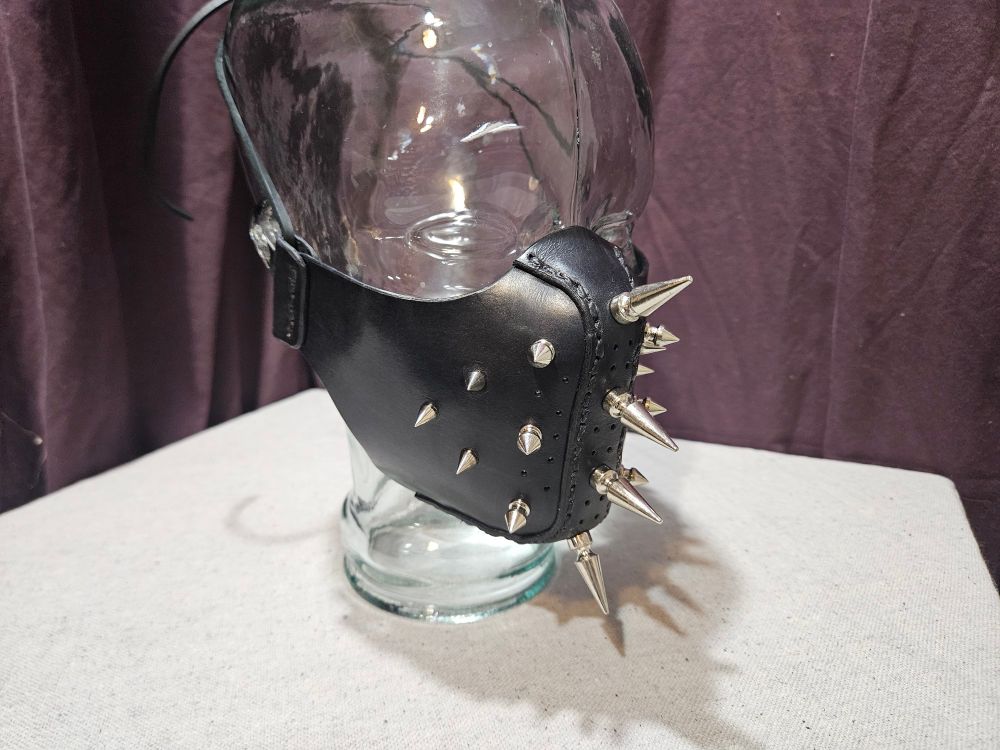 A leather half mask with polished chrome spikes in a cascading pattern of length sitting on a glass mannequin head.