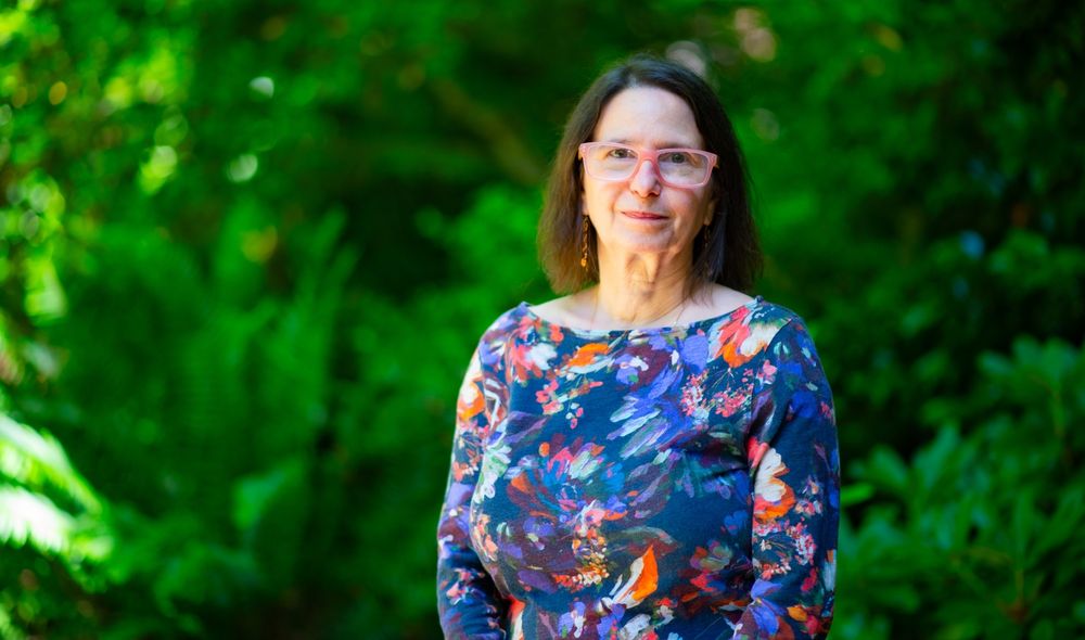 Image of Laura Schildkraut smiling, wearing a floral patterned shirt with green foliage behind her. 