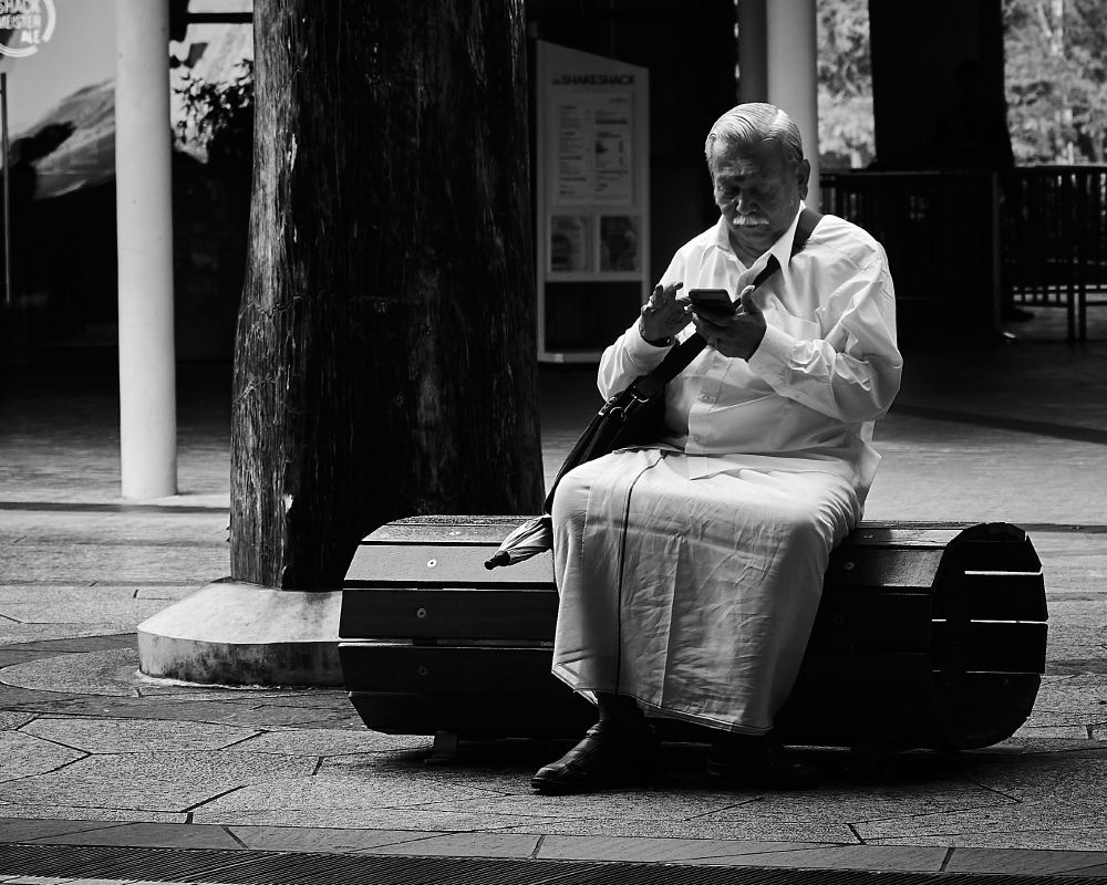An older man with short white hair sits on a wooden bench in an outdoor area. He is wearing a white shirt and light-colored pants, holding a smartphone with both hands, focused on the screen. Trees and modern structures are visible in the background