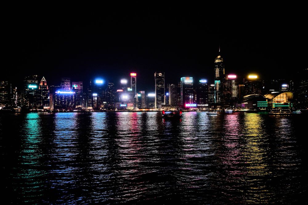 A vibrant skyline of Hong Kong Island at night. The city is filled with tall buildings illuminated with colorful lights reflecting on the calm water below, creating a shimmering display. The night sky is dark, enhancing the bright glow of the city's lights.