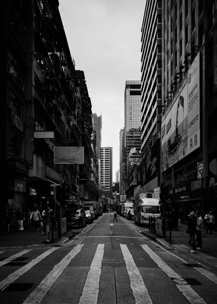 A black and white photograph of a busy urban street. Tall buildings with varied textures line both sides. The street extends into the distance, populated by people and vehicles. The scene has a strong contrast, highlighting the mix of old and new architectural styles