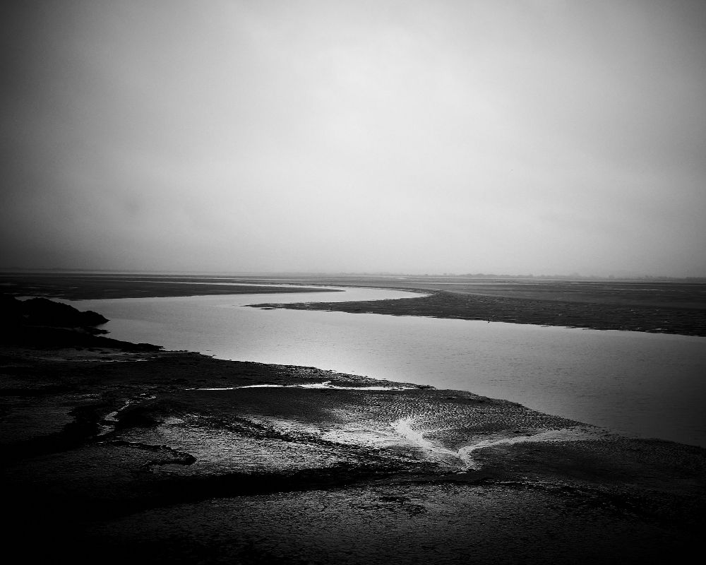 The image shows a river estuary on a cloudy day. The water reflects light, creating a shiny, winding path through dark, wet sand and mud. The sky is overcast, adding a somber atmosphere.