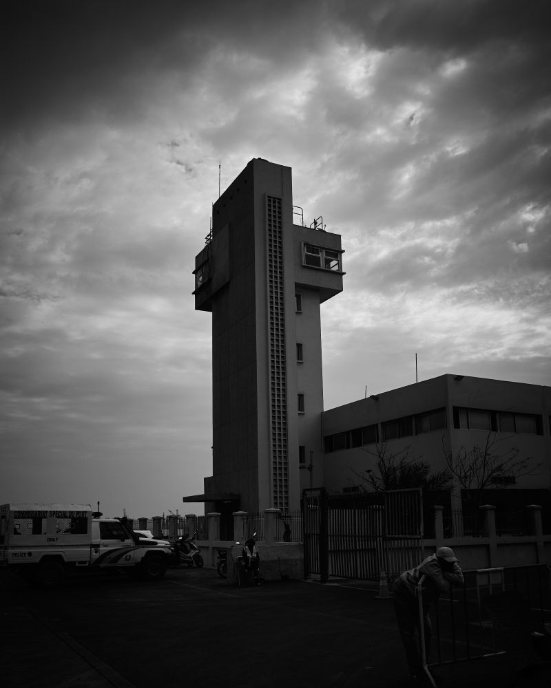 A tall control tower stands against a cloudy, overcast sky. The image is in black and white, highlighting the tower's rectangular structure and the dramatic sky