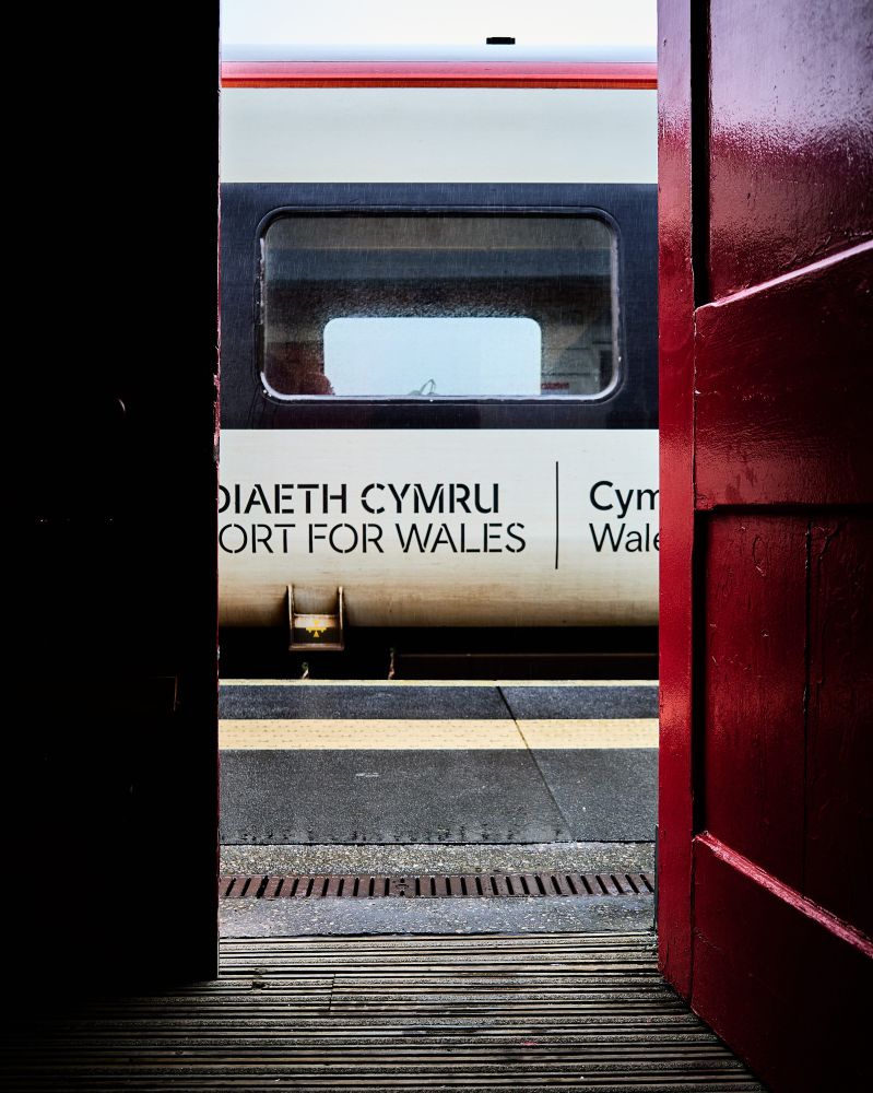 A view from an open doorway reveals a train platform and a stationary train. The train is white with a red stripe near the roof and has a window visible. The platform is textured and divided by a yellow line