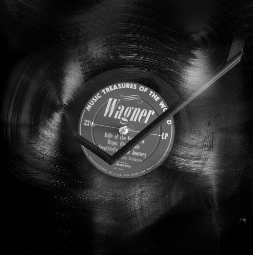 A black and white image of a vinyl record with a visible label. The label reads 'Wagner' and features a list of music tracks. The surface of the record shows scratches and light reflections, giving it a textured look. The arm of a record player is also shown