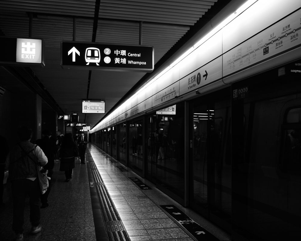 The image shows a black and white view of a subway platform in Hong Kong. People are walking along the platform, which is lined by a train with closed doors. Overhead signs display directions and station names in both English and Chinese. The station is well-lit, emphasizing the clean, organized space