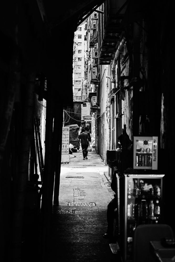 A narrow back alley in Hong Kong lined with tall buildings, with a person walking away into the distance. The alley is dark with shadows, and there is a beverage fridge on the right side