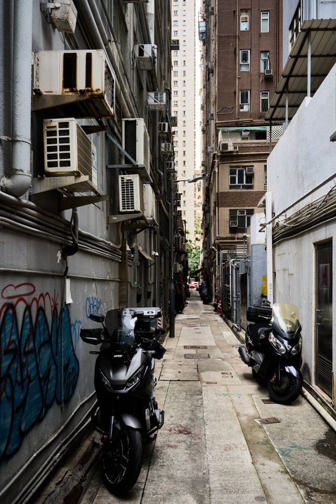 A narrow alleyway in Hong Kong lined with scooters and air conditioning units. The surrounding walls show graffiti and exposed pipes. Tall, densely packed buildings rise on either side, leading to a brighter high-rise structure in the distance