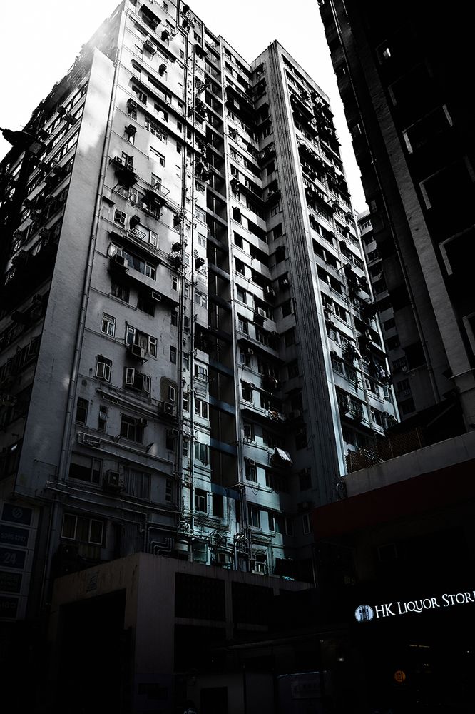 A tall, narrow building in Hong Kong rises against the sky. The exterior is marked by numerous windows and air conditioning units. Sunlight casts stark shadows, creating a pattern of light and dark across the facade