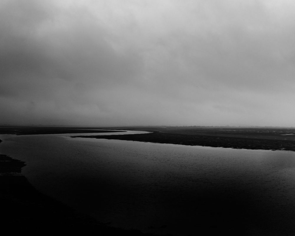 The image shows a monochrome view of a wide river estuary under a cloudy sky. The river curves gently through the landscape, leading towards a distant horizon. The scene is captured in shades of gray, conveying a calm and overcast atmosphere