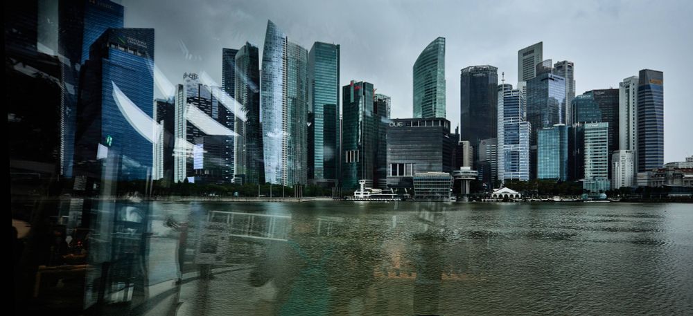 A view of a modern city skyline with tall skyscrapers. The buildings are reflected in a body of water in the foreground. The scene includes a cloudy sky, giving the image a moody atmosphere