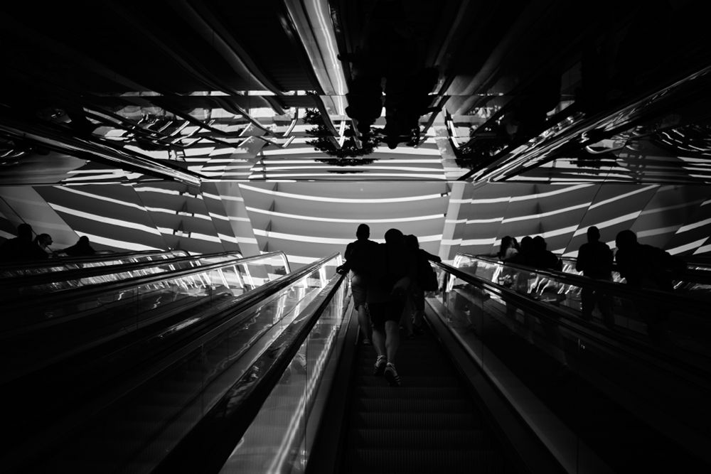 The image shows a black-and-white photo of people on an escalator. The scene is inside a modern, well-lit store with reflective surfaces and strong geometric patterns creating striking shadows.