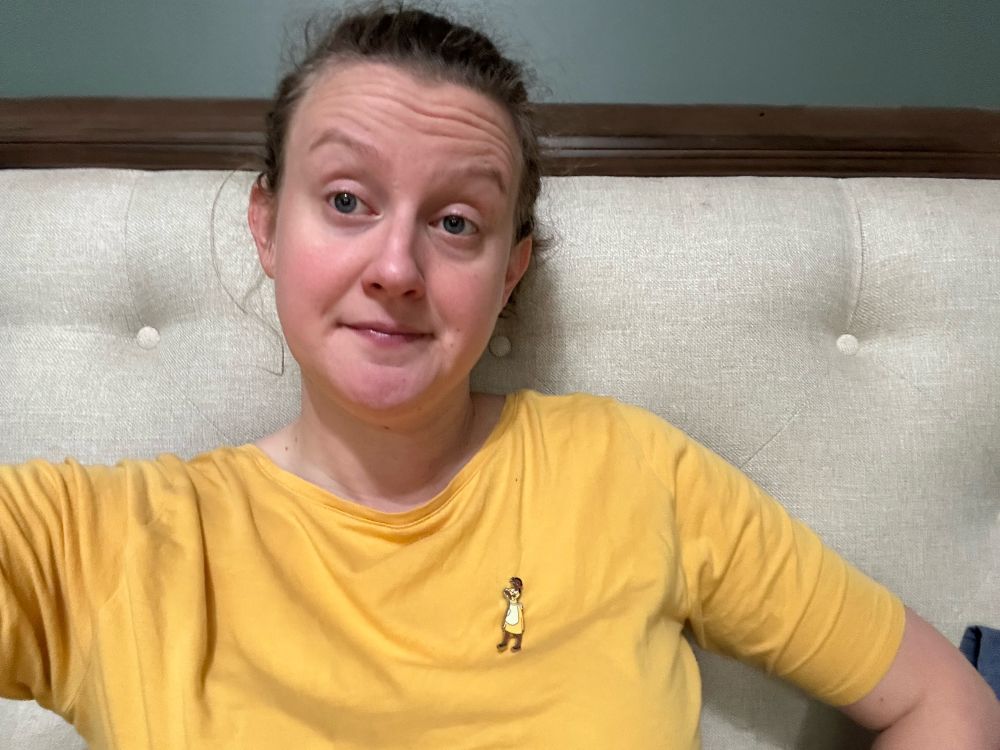 A white woman with a pony tail with her hand on her hip, wearing a yellow shirt with an enamel pin of Tiana from Princess and the Frog in her yellow waitress outfit  