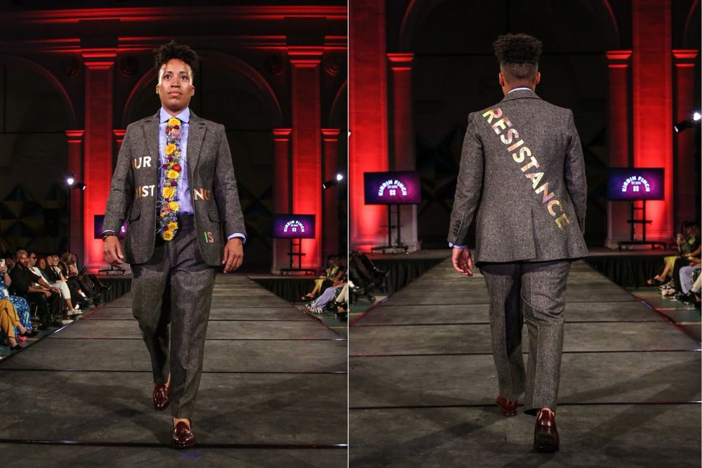 A Black model walks a runway in a Heather gray wool suit and mahogany loafers. The jacket reads “our existence is resistance.”