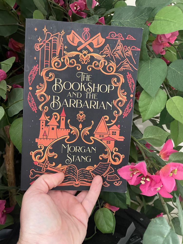 A hand holds a paperback copy of a book titled “The bookshop and the Barbarian” the book cover is black with the title and a fantasy inspired frame in shades of red and orange. The background is leafy with several artificial pink flowers. 