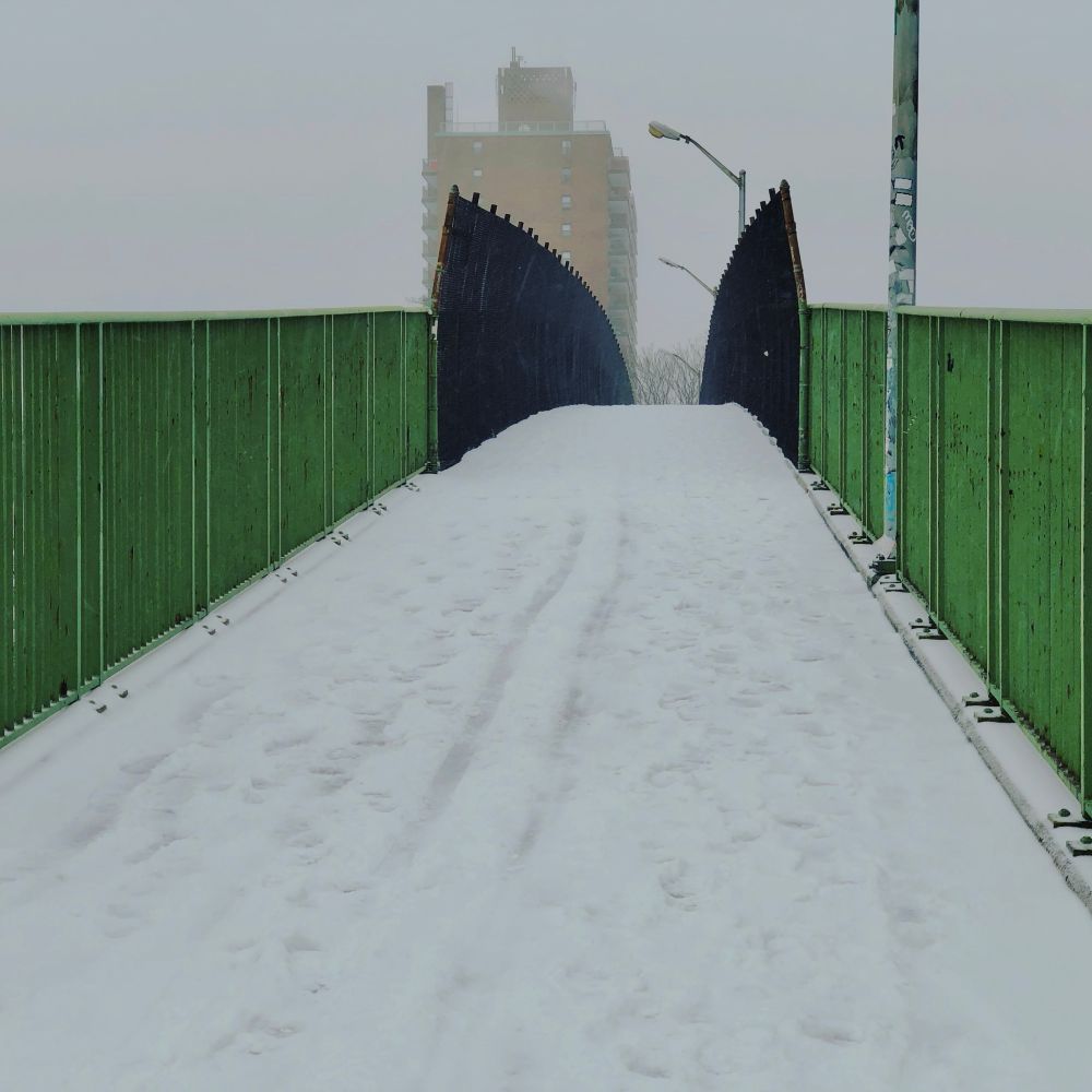 A snowy overhead pathway in the Soundview neighborhood of The Bronx sits empty. Footprints can be seen in the snow. Green painted metal fences line the sides leading up to it. A tall apartment building looms in the distance. 