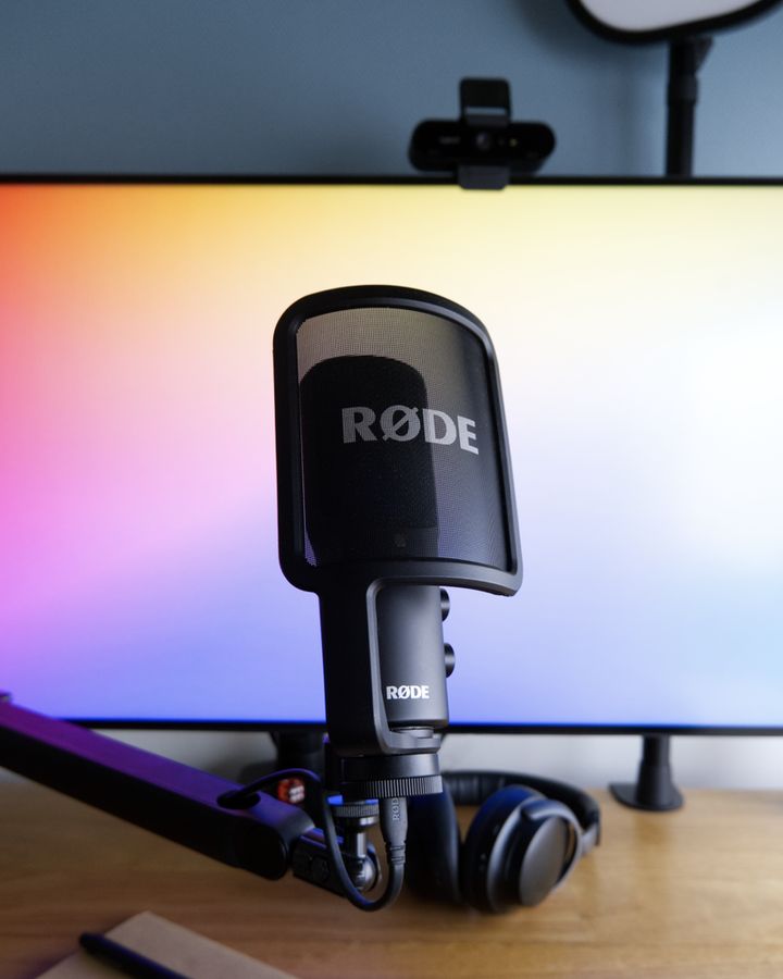 A black, desk-mounted microphone is positioned in front of a computer monitor that features a rainbow-gradient background. Also in frame are headphones, a notebook, a webcam and a desk-mounted key light.