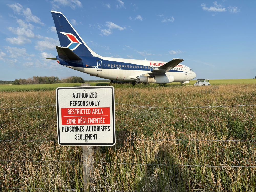 A view of the plane from a slightly different angle towards the rear. A sign is posted on the fence:

“AUTHORIZED PERSONS ONLY / RESTRICTED AREA”

Followed by the same in French:

“ZONE RÉGLEMENTÉE / PERSONNES AUTORISÉES SEULEMENT”