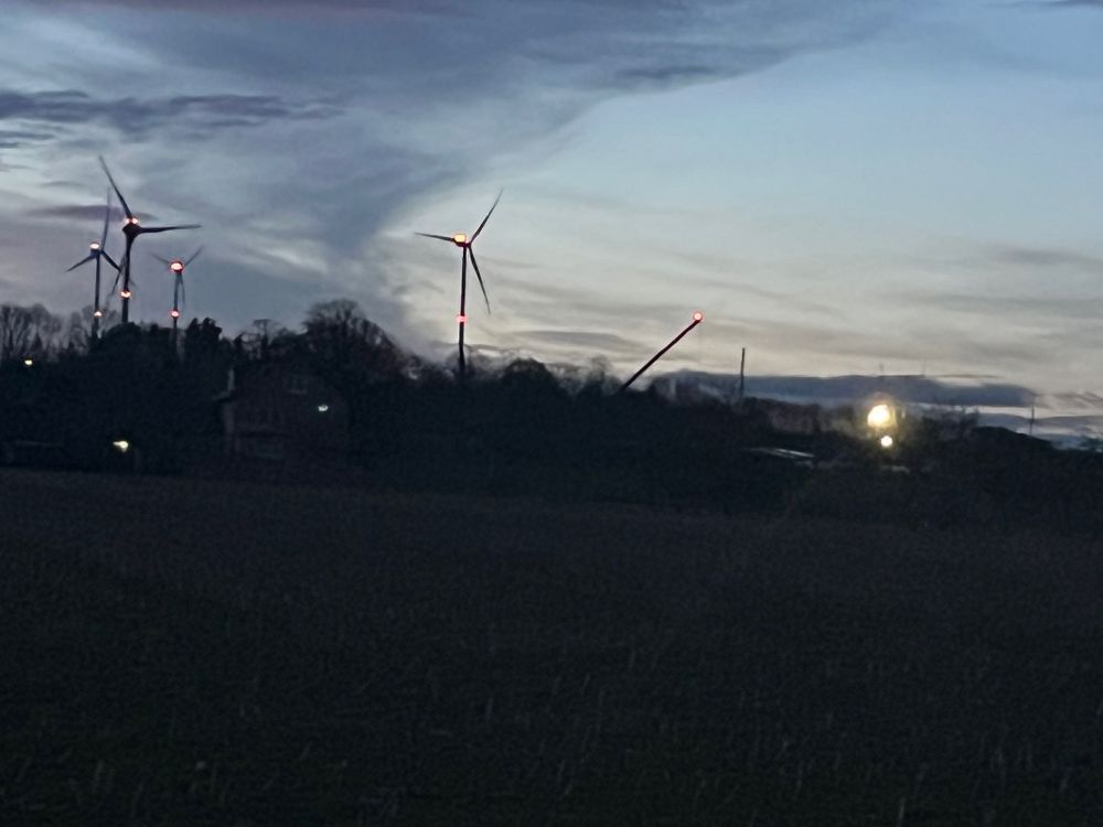 Aufziehender Morgen: die Farben wechseln von mittel- ins hellblau. Im Hintergrund blinkende Windräder. Ganz rechts darunter eines vermutlich ohne Rotorblätter und sehr schiefstehend.