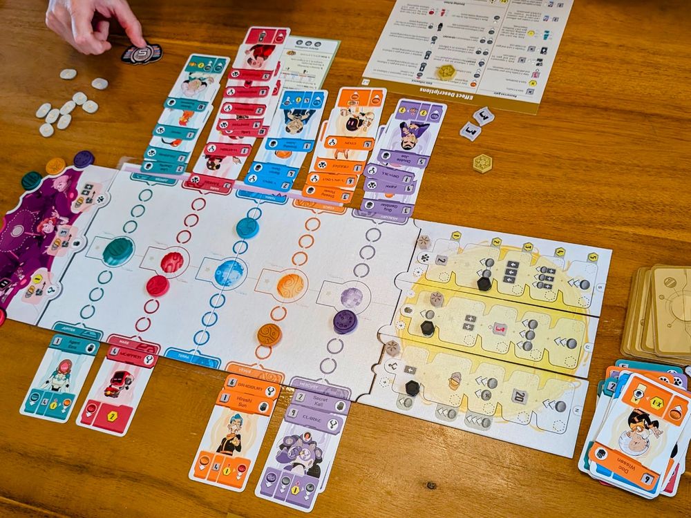 Close shot of board game in middle of play on wooden table. Cards are lined up by color in rows on each side of board and there is a scattering of bits and a hand flipping a token.