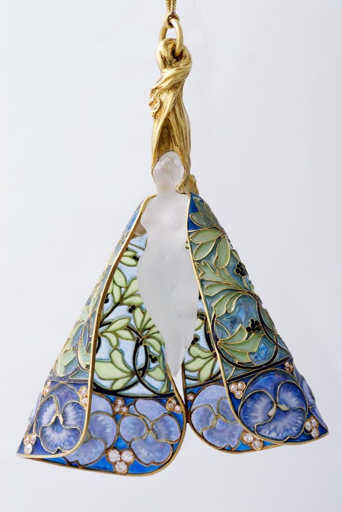 Source: "The female nude figure of the pendant is made from etched glass, and is attached to a thin chain passed through the woman’s golden cascade of hair. The shoulders of the figure are covered by a colourful cloak, the bottom of which is decorated with miniature diamonds placed in groups of three among flowers. The artistic composition is executed in a virtuoso technique, especially in the decoration of the cloak. The framework of the cloak is formed by an openwork floral pattern, leafy vines, and flowers, which are filled with greenish, bluish and violet-coloured plique-a-jour enamel. This method results in an effect similar to stained glass windows: when lit from behind, the colours of the enamel gleam. This complex technique of translucent enamel was used in the Middle Ages, but forgotten after the Renaissance. A jeweller, Andre Fernand Thesmar (1843–1912), rediscovered plique-a-jour enamel and began exhibiting objects made using this method in the 1870s. The technique became one of the hallmarks of Art Nouveau jewellery. While Thesmar used opaque enamel, Lalique (1860–1945) worked with translucent plique-a-jour enamel, recognizing its capacity to produce powerful, glowing colours. The inspiration behind the present jewel was the emblematic Parisian dancer of the period, Loie Fuller (1862–1928), whose famed choreography of 1895, the Danse serpentine, was performed in a similar, cloak-like veil."