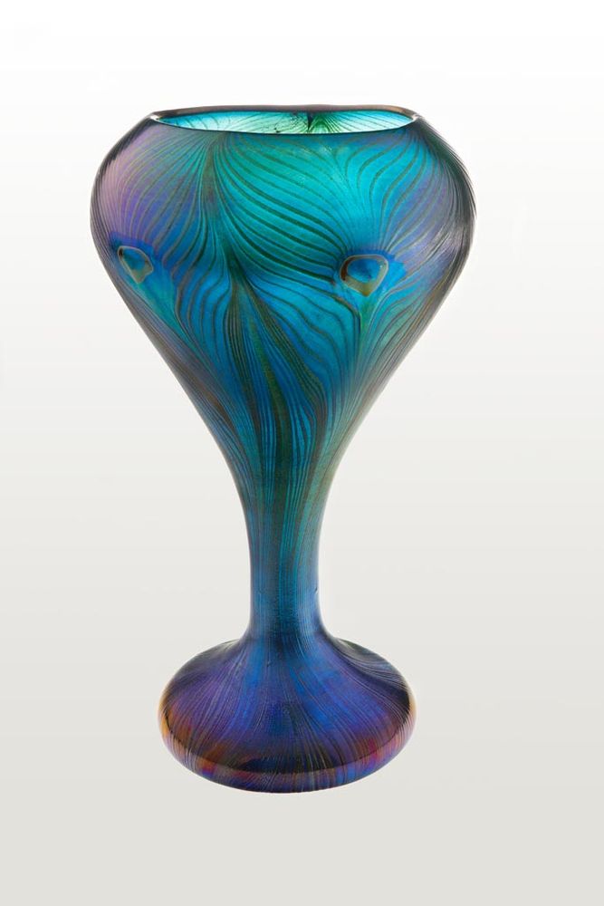 Source: "In 1898, the Director of the Museum of Applied Arts, Jenő Radisics (1856–1917) held an international exhibition entitled The Modern Art to present new developments in the contemporary art of the time. It included fifty items by Louis Comfort Tiffany (1848–1933). Prior to the Budapest exhibition, Tiffany had shown his art glass in Vienna. Siegfried Bing (1838–1905) – Tiffany’s sole distributor in Europe – wanted to avoid the embarrassment of an exhibition in Budapest featuring only pieces that Vienna customers did not want. He therefore added pieces of equivalent standard from his Paris shop La Maison de l’Art Nouveau to the Budapest exhibits. Tiffany was thus represented by a diverse set of top-quality glassware. The catalogue stated that several pieces were actually made for the exhibition. The Peacock vase was one of several other Tiffany items which the Museum of Applied Arts actually purchased. The vase displays the true shimmering blue and green tones of the bird’s feathers. The aventurine filaments placed on the blown cobalt blue base when it was still hot melted into the glass upon further heating, giving the effect of sweeping brushstrokes. The production process responsible for the Peacock vase was developed by George J. Cook, under Tiffany’s personal supervision. In an article published in 1898, Siegfried Bing wrote that Tiffany was so captivated by the peacock-feather motif that he conducted experiments for a whole year in search of a way of emulating the iridescent beauty of that magnificent bird’s plumage. His efforts gave rise to a great many Peacock vases, no two of which are alike. On the Budapest piece, the combination of refined precision with nonchalant vigour attests to virtuosity of the technique applied with a sure touch, and betrays no sign of experimental uncertainty. The vase is the embodiment of technical perfection and artistic refinement."
