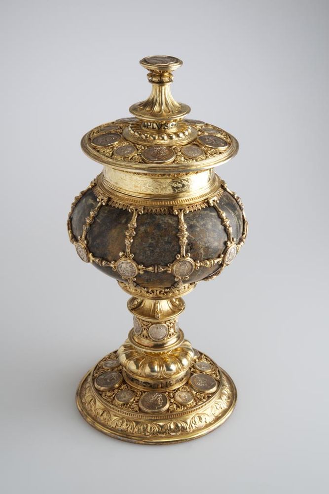 source: "The unique feature of this serpentine cup, impressive in its size, is the thirty-five various coins covering its base, bowl and lid. Most of them were made after antique originals (from the era of the Roman Empire), or are so-called fictitious coins of the Renaissance. Further interesting things to note about this goldsmith work, richly decorated with palmettos (stylised palm leaves) and acanthus tendrils are the two enamelled French inscriptions on its stem and the horizontal band of the bowl: “NVLLE PLVS” and “NE MOY AVLTRE”. These are the mottoes of Anthoine de Lalaing (1480–1540), Count of Hoogstraten (now Belgium) and his wife, Elisabeth de Coulembourg (1475–1555).
Several theories were made about the origins of this ornamental cup. According to an early idea based on the inscription of a coin, it was made in 1490 as a gift for King Matthias Corvinus. At the major goldsmith exhibition of the Museum of Applied Arts held in 1884, the work was presented as a 16th century article. Not only the date, but also the place of its manufacture is uncertain. France, Switzerland, Strasbourg, Nuremberg, and now – based on inscriptions – The Netherlands have all been considered as the place of execution of the artefact. Its precise identification requires further research."