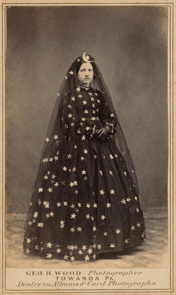 full length portrait of a woman in 1860s style dress, but the dress is dark with white stars covering it, She also has a long veil with white stars, and a headdress with a moon on it