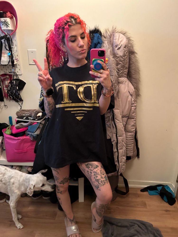 This is a mirror selfie. Fay is wearing an oversized black t-shirt by the Toronto rapper DT. She has her pink and orange hair in a high pony and is making a piece sign with one hand while holding her phone on the other.  Her many tattoos are visible in her arms and legs and her small-to-medium white dog is seen sniffing her knee. 