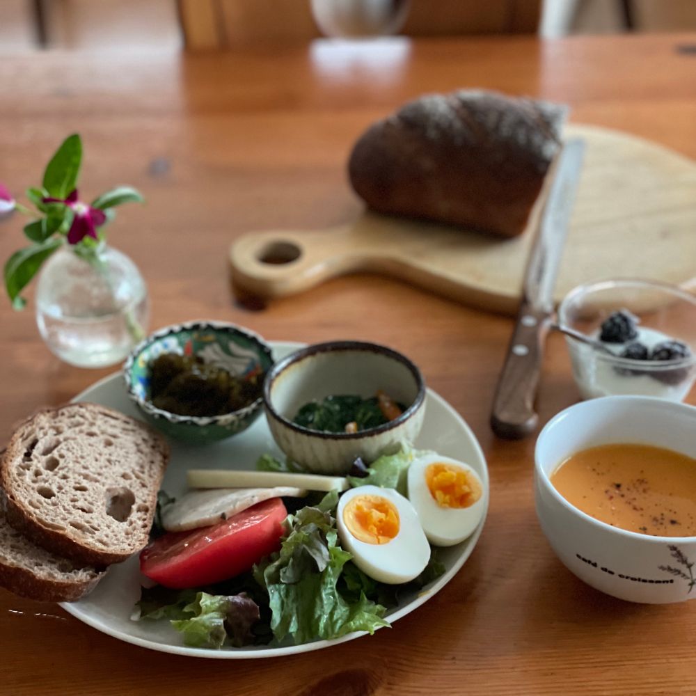 朝食　白丸皿に天然酵母全粒粉100%パン ゆで卵 サニーレタス トマト ハム ゴーダチーズ 小鉢のもずく・モロヘイヤ人参胡麻和えが載っている　冷製人参ポタージュ　低脂肪乳ヨーグルト冷凍ブラックベリー添え　奥のカットボードに切りかけのパン