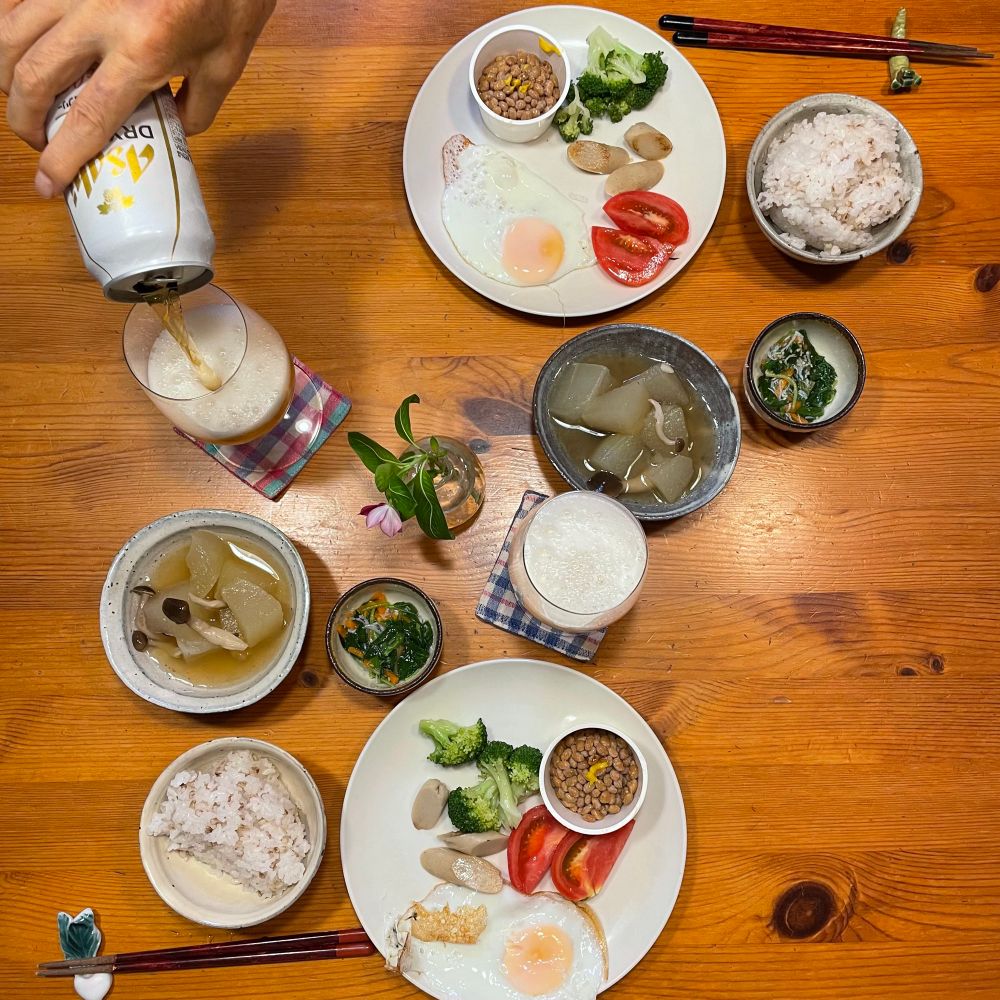 2人分の夕食テーブルでノンアルビールを注いでいる　目玉焼き魚肉ソーセージとブロッコリー炒めトマト納豆が白丸皿に載っている　冬瓜しめじ炊いたん　モロヘイヤ人参胡麻じゃこ和え　もち麦ごはん