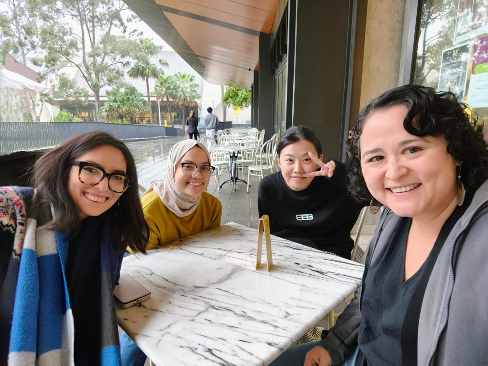 Four female scientists smiling and looking at the camera while sitting in a cafe. A building, cafe tables and trees are visible in the background.