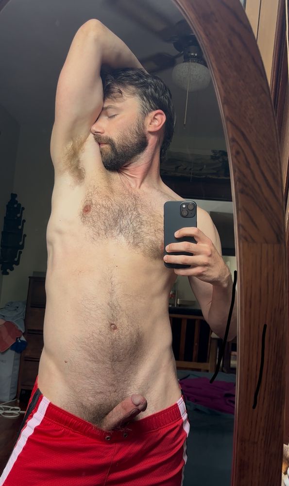 Mirror selfie of Patrick with one arm over his head and his face in his hairy pit. He’s wearing red shorts and his hard dick is popping out of the top