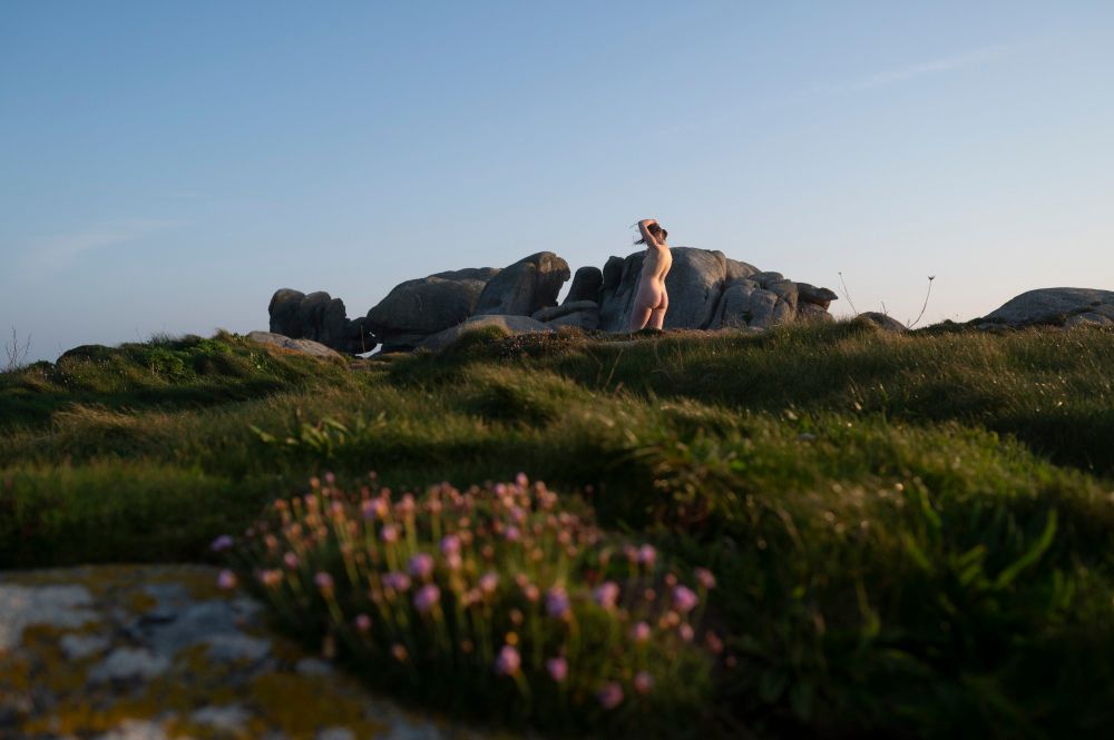 Naked woman from back posing in front of big granite rocks, in the foreground an armeria maritima bush catches sun light.