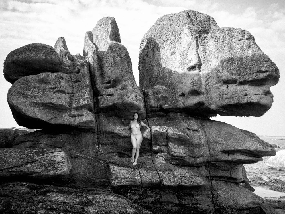 Naked woman standing in front of a big rock. She watches the viewer, both arm open du give her some grip on the rock. The picture is black and white.
