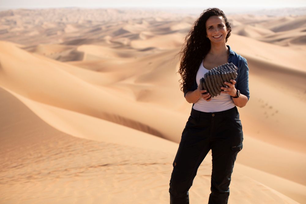 Presenter Ella Al-Shamahi standing in a desert holding an object.