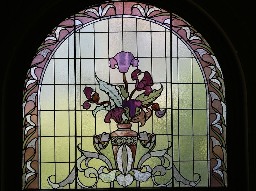 Ein rundbogenfenster mit rosafarbener Glas-Randung. IN der Mitte des Fenster ist eine Vase mit lila Iris zu sehen. 