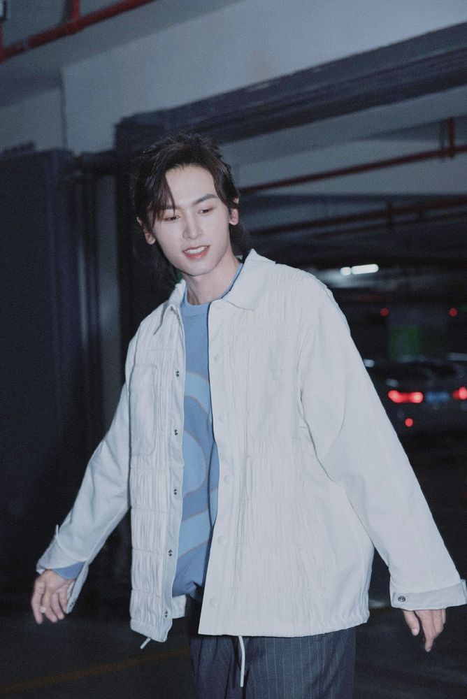 Chinese actor Zhang Zhehan for a photoshoot in 2020, here his hair are tied up, he's wearing a white a very light white shirt with another blue shirt underneath. In this picture he's smiling 